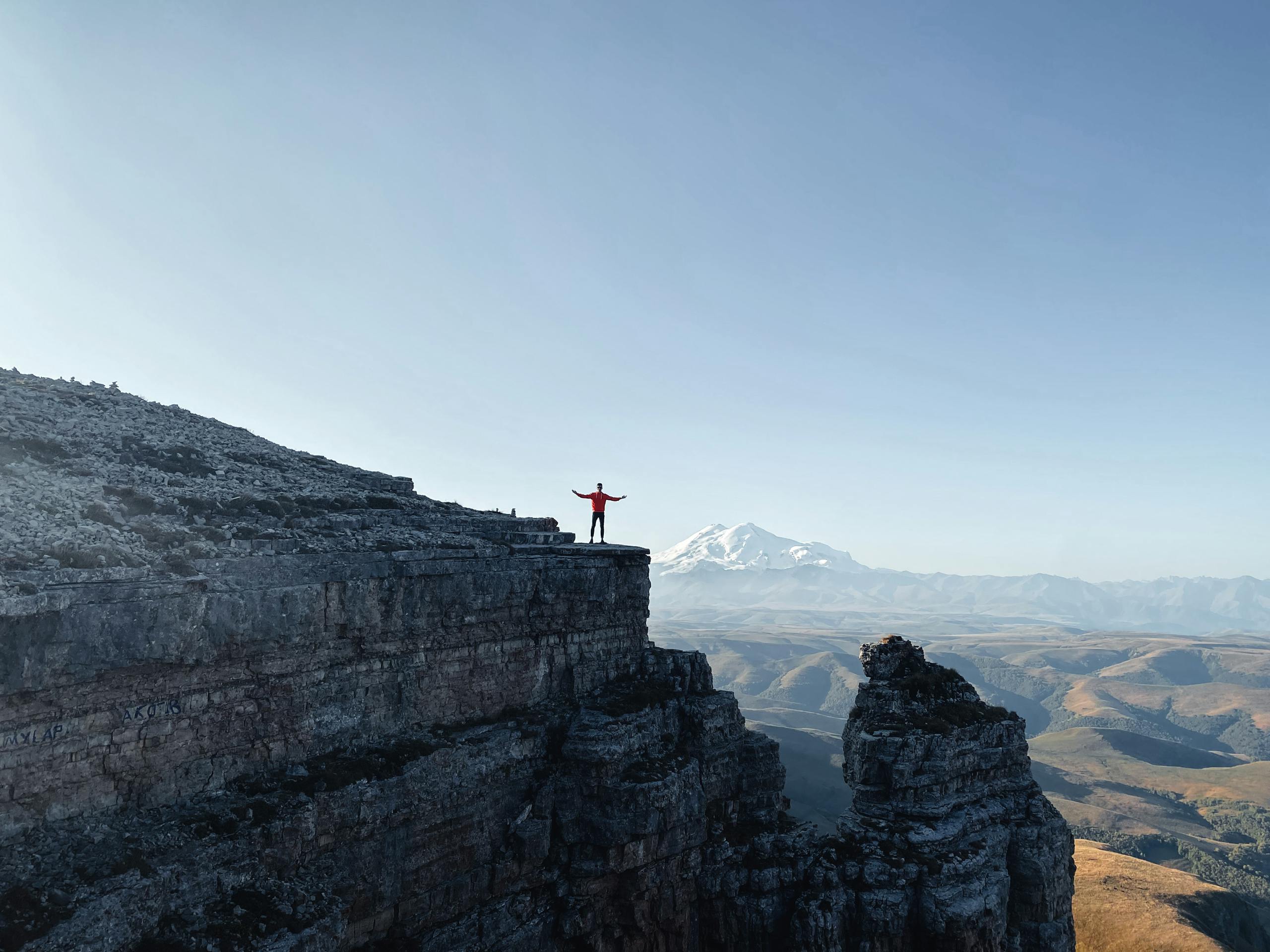 A lone person stands triumphantly on a cliff, with Mount Elbrus visible in the distance under a clear sky.